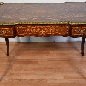 May include: A rectangular antique desk with a dark wood finish and intricate inlaid floral patterns. The desk features three drawers with floral marquetry and ornate brass accents on the corners and legs. The legs are curved.