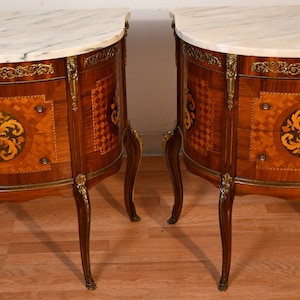 May include: A pair of semi-circular wooden cabinets featuring white marble tops with grey veining. The front panels display intricate marquetry with geometric patterns and central floral motifs, accented by ornate gold-toned metal mounts and legs.