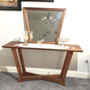 Puede incluir: Una consola de madera con espejo incorporado. La mesa tiene una parte superior rectangular y patas anguladas. Un camino de mesa de encaje blanco y objetos decorativos se encuentran sobre la mesa. El espejo está enmarcado en madera y se apoya en la mesa.