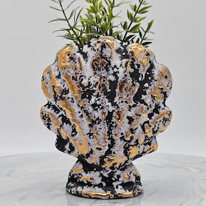 May include: A decorative seashell-shaped vase with a black, white, and gold textured finish. The vase holds green leafy stems. The vase has a pedestal base and is suitable for home decor.