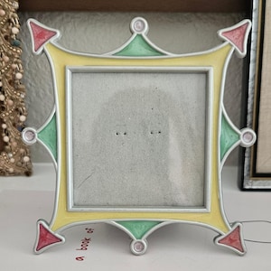 May include: A square picture frame with a yellow border, accented with red and green arrow-shaped details. The frame has a silver trim and small pink circular embellishments. The frame is empty, ready for a photo.