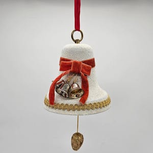 May include: A white, glitter-covered bell-shaped Christmas ornament with a red velvet bow and gold trim. Two small silver bells hang below the bow. A gold-colored, teardrop-shaped ornament dangles from the bottom, suspended by a gold string.