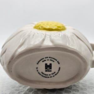 Peut inclure: Mug en c&eacute;ramique blanche en forme de marguerite avec un centre jaune. Le mug a une anse et est marqu&eacute; "Dishwasher and Microwave Safe Made in China" en bas. Un charmant design floral pour les articles de cuisine.