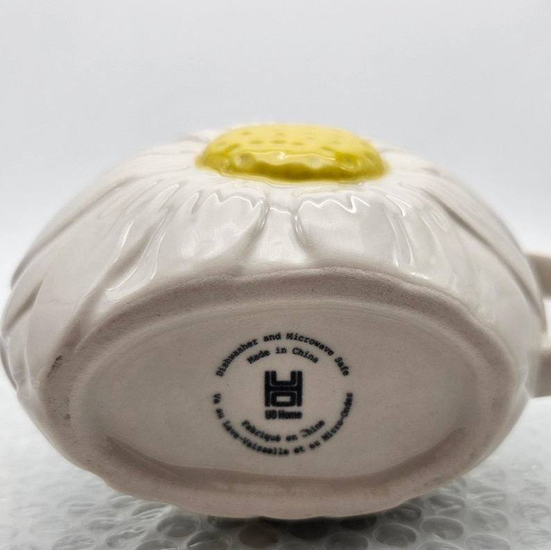 Peut inclure: Mug en c&eacute;ramique blanche en forme de marguerite avec un centre jaune. Le mug a une anse et est marqu&eacute; "Dishwasher and Microwave Safe Made in China" en bas. Un charmant design floral pour les articles de cuisine.
