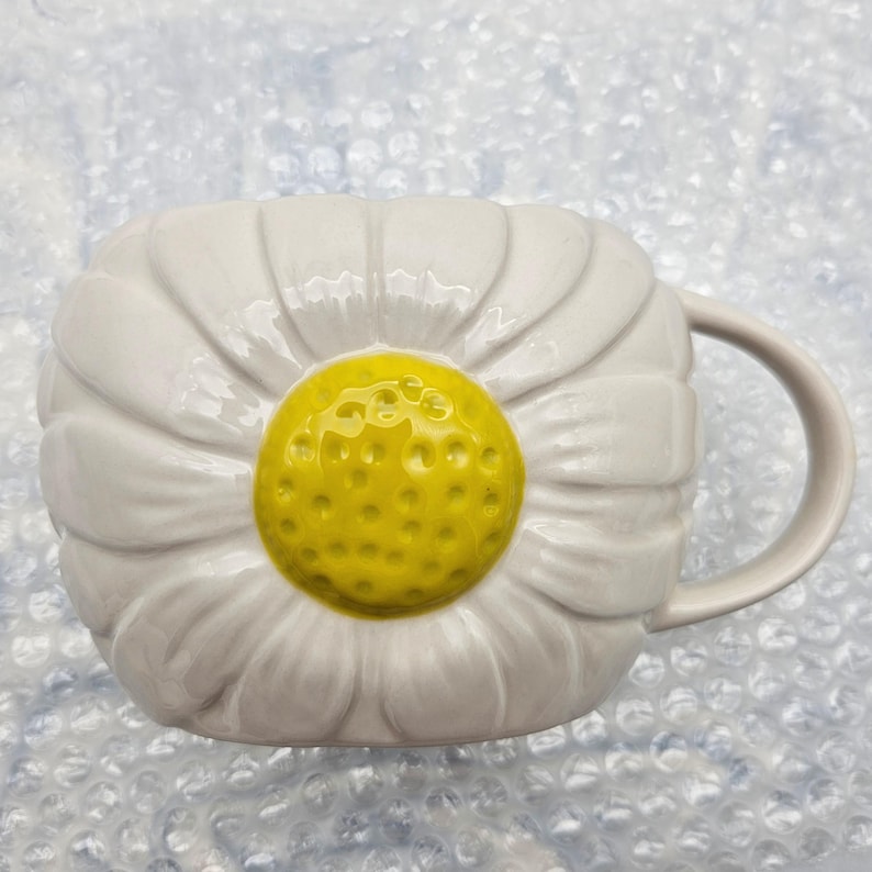 Peut inclure: Une tasse en c&eacute;ramique blanche en forme de marguerite, avec un centre jaune et des p&eacute;tales blancs. La tasse a une poign&eacute;e incurv&eacute;e et est con&ccedil;ue pour ressembler &agrave; une fleur. Un article unique et d&eacute;coratif pour savourer des boissons.
