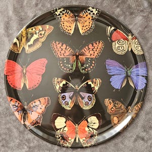 May include: A round, black serving tray featuring a collection of colorful butterfly illustrations. The butterflies are in various colors, including orange, red, blue, and yellow, with detailed wing patterns. The tray has a glossy finish.