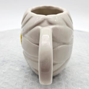 Peut inclure: Une tasse en c&eacute;ramique cr&egrave;me avec un motif de momie. La tasse pr&eacute;sente un motif enveloppant de bandages et une anse incurv&eacute;e. Un petit accent jaune est visible sur le c&ocirc;t&eacute;. La tasse est con&ccedil;ue pour les boissons.