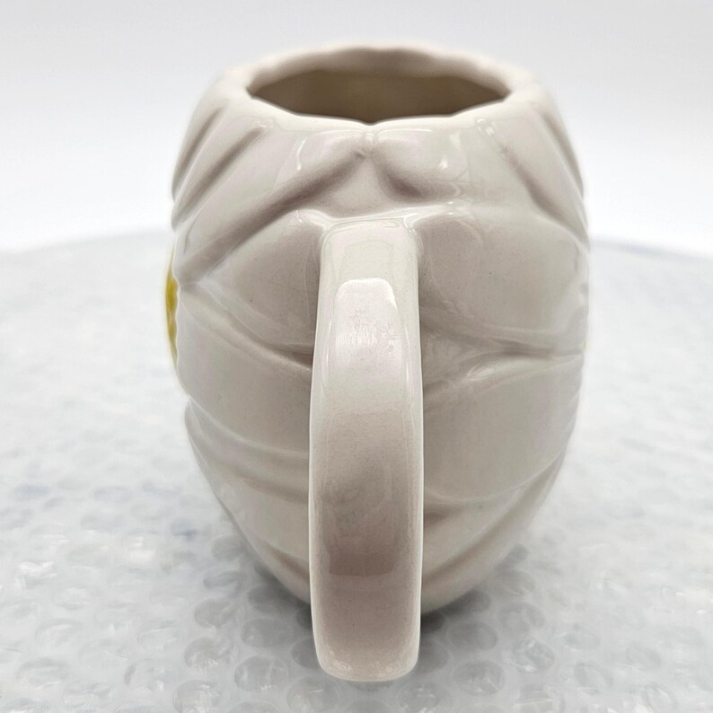 Peut inclure: Une tasse en c&eacute;ramique cr&egrave;me avec un motif de momie. La tasse pr&eacute;sente un motif enveloppant de bandages et une anse incurv&eacute;e. Un petit accent jaune est visible sur le c&ocirc;t&eacute;. La tasse est con&ccedil;ue pour les boissons.