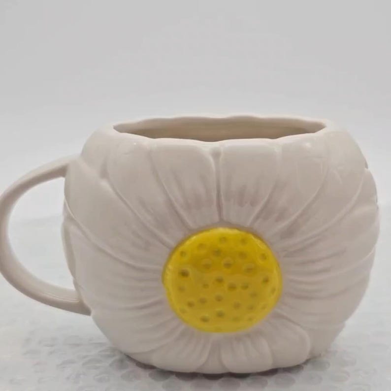 Peut inclure: Une tasse en c&eacute;ramique en forme de fleur de marguerite. La tasse est blanche avec des d&eacute;tails de p&eacute;tales et un centre jaune. La poign&eacute;e est incurv&eacute;e et blanche, et l'int&eacute;rieur est beige clair.