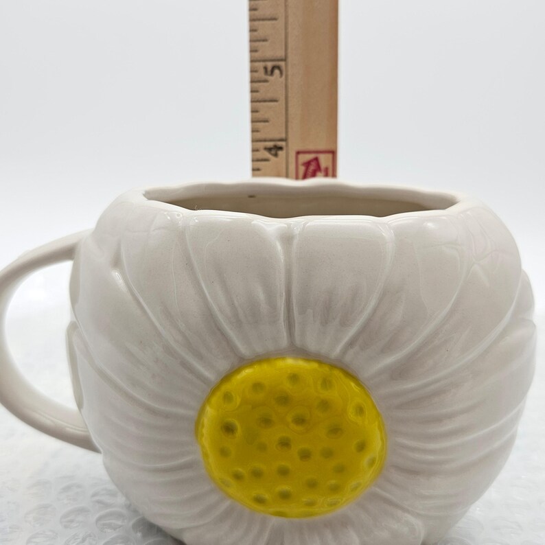 Peut inclure: Mug en c&eacute;ramique blanche en forme de fleur de marguerite, avec un centre jaune et une anse blanche. Le mug mesure environ 10 cm de haut. Le mug est con&ccedil;u pour ressembler &agrave; une fleur de marguerite.