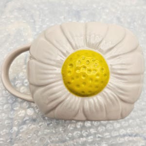 Peut inclure: Une tasse en c&eacute;ramique blanche en forme de fleur de marguerite. La tasse pr&eacute;sente un centre jaune et des p&eacute;tales blancs, avec une anse incurv&eacute;e sur le c&ocirc;t&eacute;. Le design est en trois dimensions, ce qui donne &agrave; la tasse une apparence florale unique.