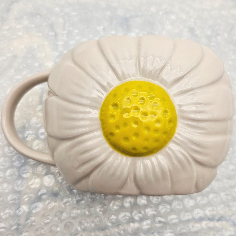 Peut inclure: Une tasse en c&eacute;ramique blanche en forme de fleur de marguerite. La tasse pr&eacute;sente un centre jaune et des p&eacute;tales blancs, avec une anse incurv&eacute;e sur le c&ocirc;t&eacute;. Le design est en trois dimensions, ce qui donne &agrave; la tasse une apparence florale unique.