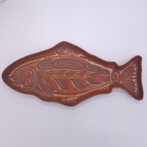 May include: A brown ceramic serving platter in the shape of a fish. The platter features a detailed, carved design of a fish with intricate patterns. The fish-shaped dish is a decorative piece for serving food.