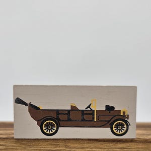 May include: A brown vintage car illustration with black and yellow accents on a rectangular wooden block. The car features a black cat on the back and detailed wheels. The background is a neutral color.
