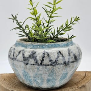 May include: A small, round ceramic planter with a distressed, textured finish. The planter features a blue and white color scheme with a geometric pattern. Green leafy plant stems sprout from the top of the planter, adding a natural element.