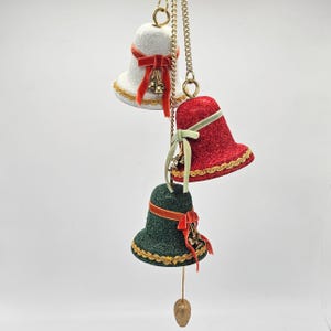 May include: Three hanging Christmas bell ornaments. The bells are white, red, and green, with gold trim and red velvet bows. Each bell has a small cluster of gold bells and hangs from a gold chain. A gold teardrop-shaped ornament hangs from the bottom bell.