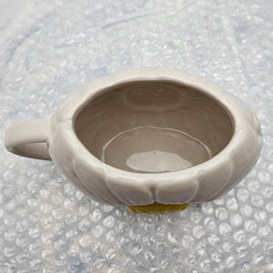 Peut inclure: Une tasse en c&eacute;ramique blanche avec un motif en forme de p&eacute;tales sur le bord. La tasse a une petite anse et une base jaune. L'int&eacute;rieur est de couleur beige clair. La tasse est probablement utilis&eacute;e pour boire des boissons.