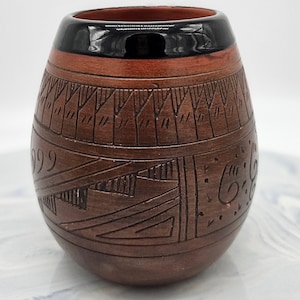 May include: A rounded brown clay vessel featuring a smooth, glossy black rim. The exterior surface is decorated with intricate, hand-carved geometric patterns, including triangular motifs and swirling lines, etched directly into the terracotta-colored clay.