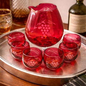 May include: A ruby red glass pitcher and matching glasses set on a silver tray. The pitcher and glasses feature a grape vine design. The set is displayed on a wooden surface, suggesting a vintage or antique aesthetic.