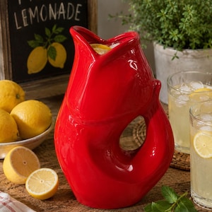May include: A bright red ceramic pitcher shaped like a fish with an open mouth at the top and a handle formed by the tail. The pitcher is filled with a beverage containing ice and lemon slices, featuring a glossy finish and a curved body design.