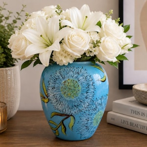May include: A bright blue ceramic vase featuring hand-painted dark blue and green floral motifs, filled with a lush arrangement of white lilies, roses, and small filler flowers. The vase has a rounded shape and a glossy finish, showcasing detailed brushwork.