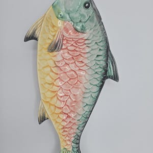 May include: A ceramic fish-shaped platter with a colorful design. The head is green, the body is yellow and pink, and the tail is green. The fish has black fins and a detailed scale pattern.