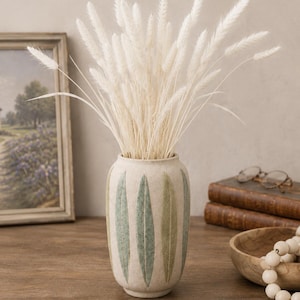 May include: A cylindrical ceramic vase with a textured off-white finish and vertical green leaf-shaped patterns. The vase is filled with a bouquet of tall, fluffy, white dried grass plumes that extend upwards and outwards from the opening.