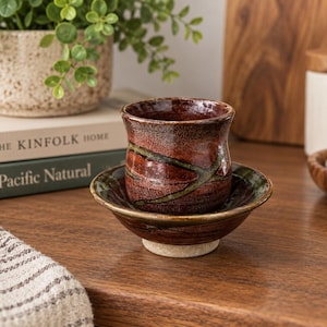 May include: A small, cylindrical ceramic cup with a glossy, mottled reddish-brown glaze and a dark green diagonal stripe, resting inside a matching shallow ceramic saucer with a flared rim and a light-colored, unglazed circular base.
