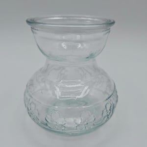 May include: A clear glass vase with a bulbous base and a smaller, rounded top. The base features a floral pattern, and the vase has a unique hourglass shape. The transparent design allows light to pass through, highlighting the details.