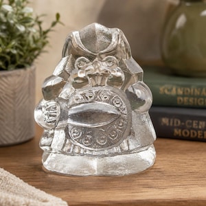 May include: A clear, decorative figurine of a seated figure with a detailed design. The figure has a round shield with an ornate pattern. The figurine is on a wooden surface, with a potted plant and books in the background.