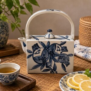 May include: A rectangular ceramic teapot with a high arched handle and a small lid, featuring a hand-painted blue floral pattern on a white background. The teapot has a short spout on the left side and a flat base, showcasing a classic blue and white design.