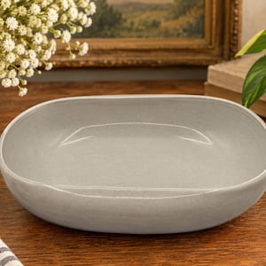 May include: A shallow, oval-shaped ceramic bowl with a smooth, light gray matte finish. The bowl features a wide, open interior and a slightly curved rim, presenting a clean and simple design suitable for serving food or as a decorative piece.
