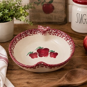 May include: A cream-colored, apple-shaped ceramic bowl with a red speckled rim and apple illustrations. The bowl has a heart-shaped handle and sits on a wooden surface. A jar labeled "SUGAR" is in the background.
