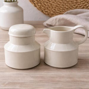 May include: Cream-colored ceramic sugar bowl with lid and a matching creamer pitcher. The sugar bowl is cylindrical with a lid, and the creamer has a handle and spout. Both are on a light wood surface.