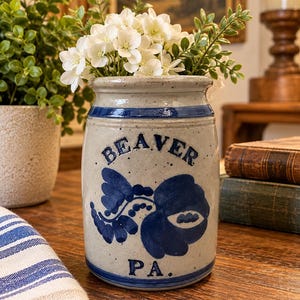 May include: A cylindrical stoneware crock with a speckled light gray finish and blue painted accents. The front features the text BEAVER and PA in blue, separated by a blue floral motif. A blue band encircles the top and bottom of the vessel.