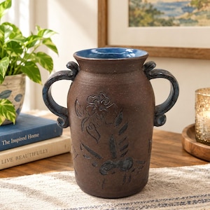 May include: A dark brown ceramic vase with two curved handles and a vibrant blue interior. The exterior features a subtle, embossed floral pattern. The vase has a wide, flared rim and a sturdy, cylindrical body with a textured, matte finish.