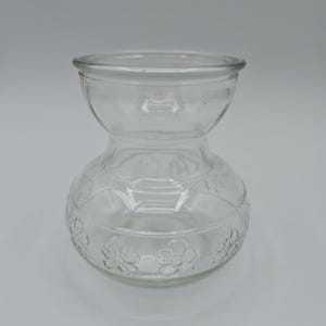 May include: A clear glass vase with a bulbous base and a smaller, flared top. The base features a repeating floral pattern. The vase is empty and sits against a plain background.