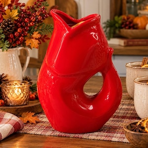 May include: A bright red ceramic pitcher shaped like a fish with an open mouth at the top and a curved tail forming the handle. The glossy, solid-colored vessel features a smooth, rounded body and a distinct gill detail on the side.
