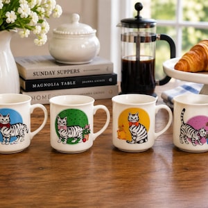 May include: Four white ceramic mugs featuring illustrations of a grey tabby cat. Each mug displays the cat against a different colored circular background: blue, green, yellow, and pink. The cat wears a bow tie in each scene with small accessories.