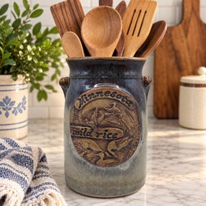 May include: A cylindrical ceramic utensil crock with a blue and grey glazed finish. The front features a raised circular medallion with the embossed text Minnesota wild rice and a detailed relief of flying ducks in a marshy landscape.