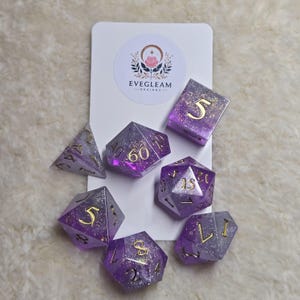 May include: A collection of seven purple and silver polyhedral dice with gold numerals. The dice are displayed on a white card featuring the "EVEGLEAM DESIGNS" logo, with a floral emblem.