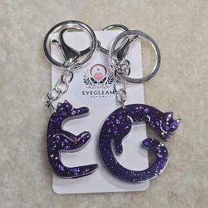 May include: Two silver keychains with purple glitter cat charms. The charms are shaped like the letters E and G. The keychains are attached to the charms with silver rings. The keychains are labelled "Evegleam Designs".