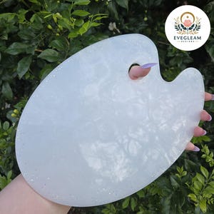 May include: A white, oval artist's palette with a pearlescent finish and glitter accents. The palette has a thumb hole. The background is green foliage. The logo "EVEGLEAM DESIGNS" is visible in the upper right corner.
