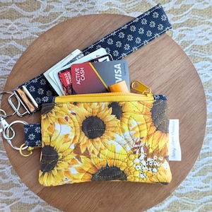 May include: A small, yellow sunflower-patterned wallet with a gold zipper, a navy blue wrist strap, and a navy blue key fob. The wallet contains credit cards and cash. The wrist strap and key fob have a white floral pattern.