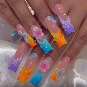 May include: Long, square-shaped acrylic nails with a gradient design in shades of orange, yellow, blue, and purple. Each nail features a 3D flower or bow accent. The nails have a clear base with a French tip.