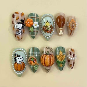 May include: A set of ten artificial nails with fall-themed designs. The nails feature Snoopy, pumpkins, leaves, and other autumn motifs. Colors include orange, green, brown, and beige, with pearl accents.