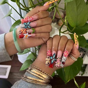 May include: Long, square-shaped acrylic nails with intricate 3D floral and animal print designs. The nails feature a variety of colors, including pink, red, blue, and white, with accents of gold and black. Some nails have a leopard print design.