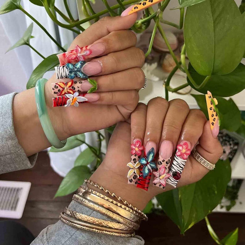 May include: Long, square-shaped acrylic nails with intricate 3D floral and animal print designs. The nails feature a variety of colors, including pink, red, blue, and white, with accents of gold and black. Some nails have a leopard print design.