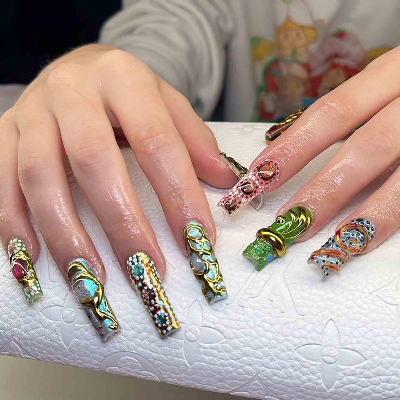 May include: A hand with long, decorated fingernails. The nails feature various designs, including gold embellishments, gemstones, and intricate patterns. Colors include gold, red, green, blue, and brown. The nails are on a white surface.