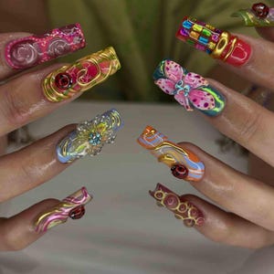 May include: A close-up of several long, sculpted acrylic nails. Each nail features a unique design, including floral embellishments, colorful patterns, and 3D elements. The nails showcase a variety of colors, including pink, gold, blue, and orange.
